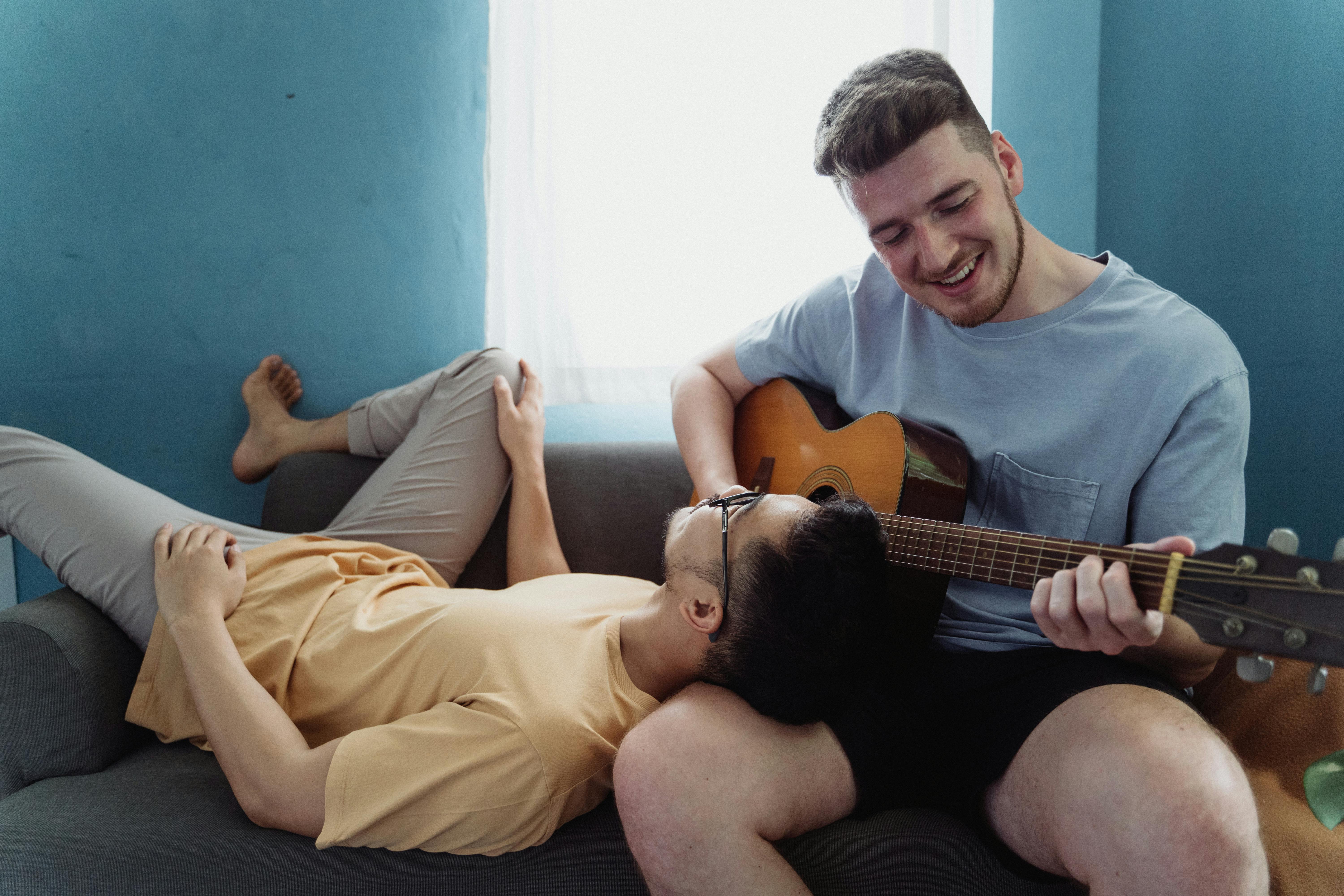 man playing guitar while another man rests his head on his leg