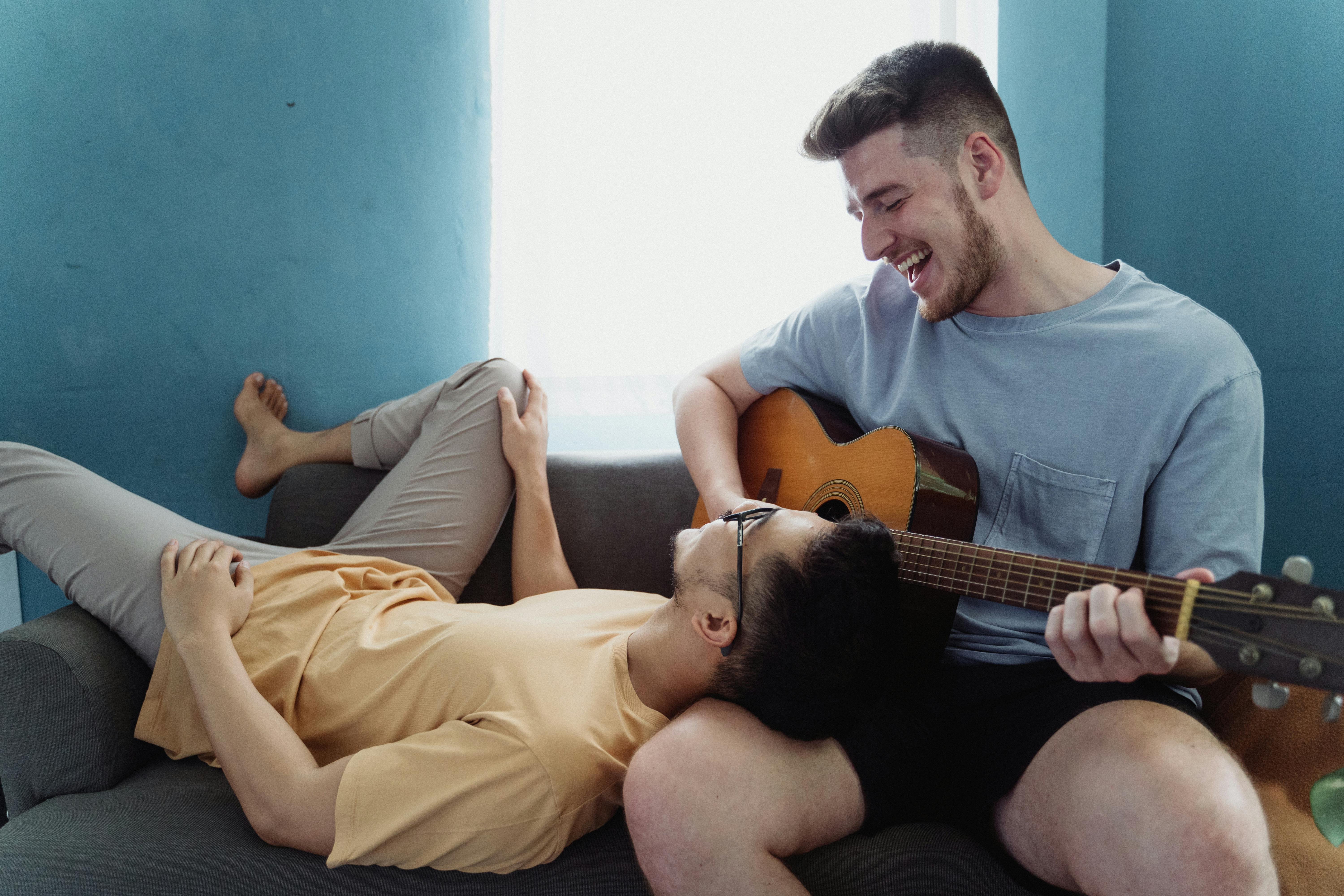 man playing guitar while another man rests his head on his leg