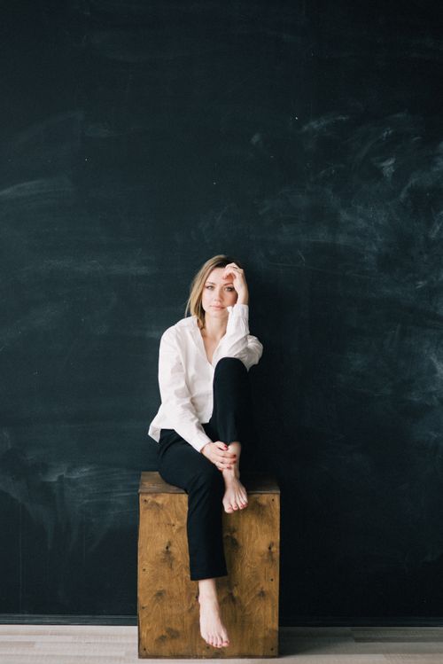 Free Elegant woman in black pants and white shirt sitting barefoot on a wooden box indoors. Stock Photo