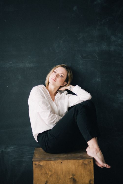 Free Elegant portrait of a woman in black pants and white shirt seated on a wooden box against a dark backdrop. Stock Photo