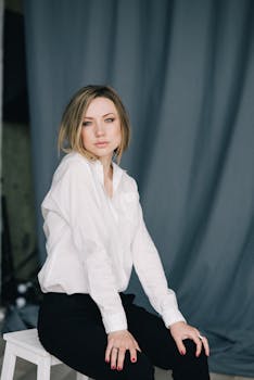 Stylish portrait of a woman sitting indoors in a white shirt and black pants against a neutral backdrop.