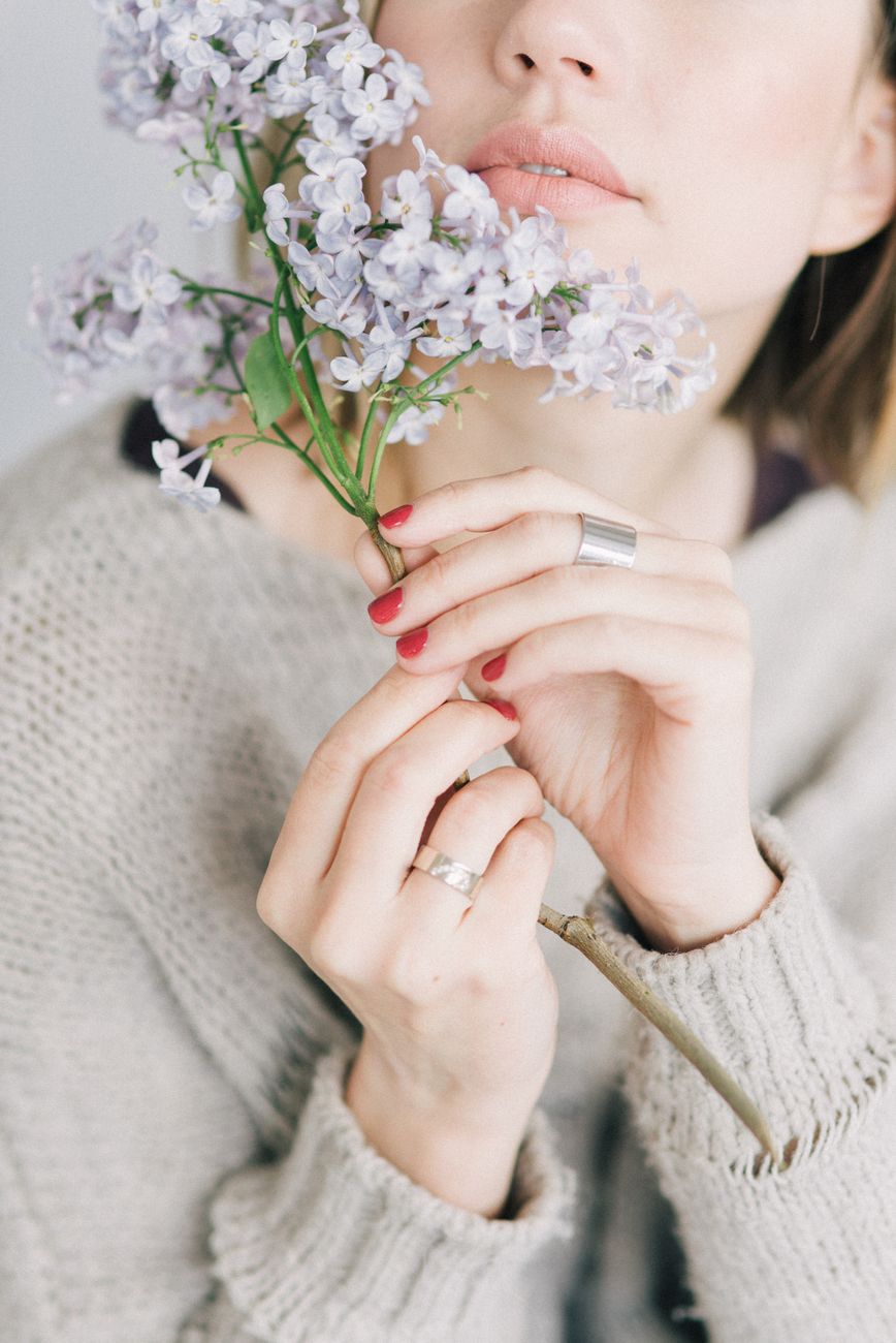 Close-up portrait of a woman with red nails, holding delicate lilac flowers, wearing a cozy sweater.