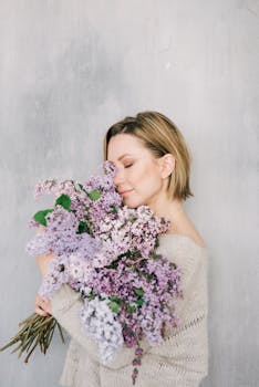 Blonde woman with short hair embraces lilacs, eyes closed, conveying tranquility.