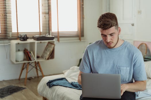 A bearded man in a blue t-shirt focused on a laptop in a sunlit bedroom.