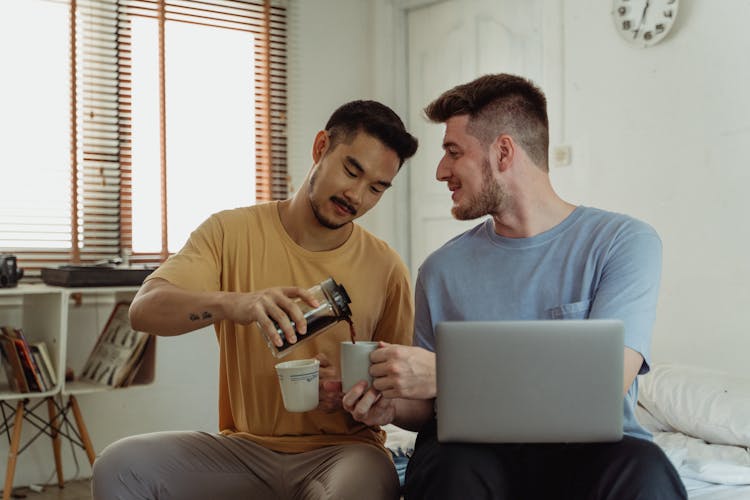 Men Pouring Coffee Into Mugs, Sitting In Bedroom And Using Laptop