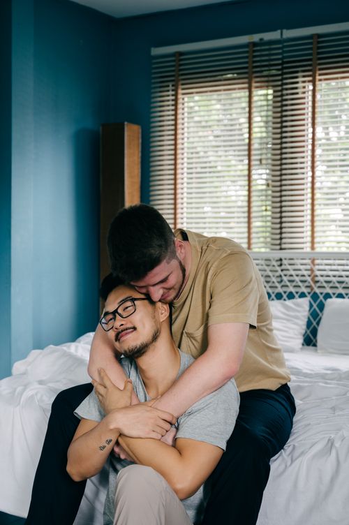 Free An interracial gay couple sharing an intimate moment while sitting on a bed in a cozy room. Stock Photo