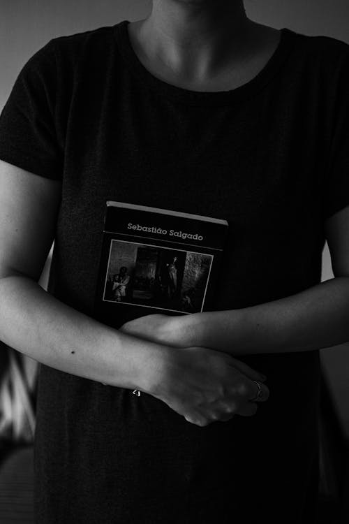 Black and white of crop anonymous young female standing and crossing hands holding book with black cover