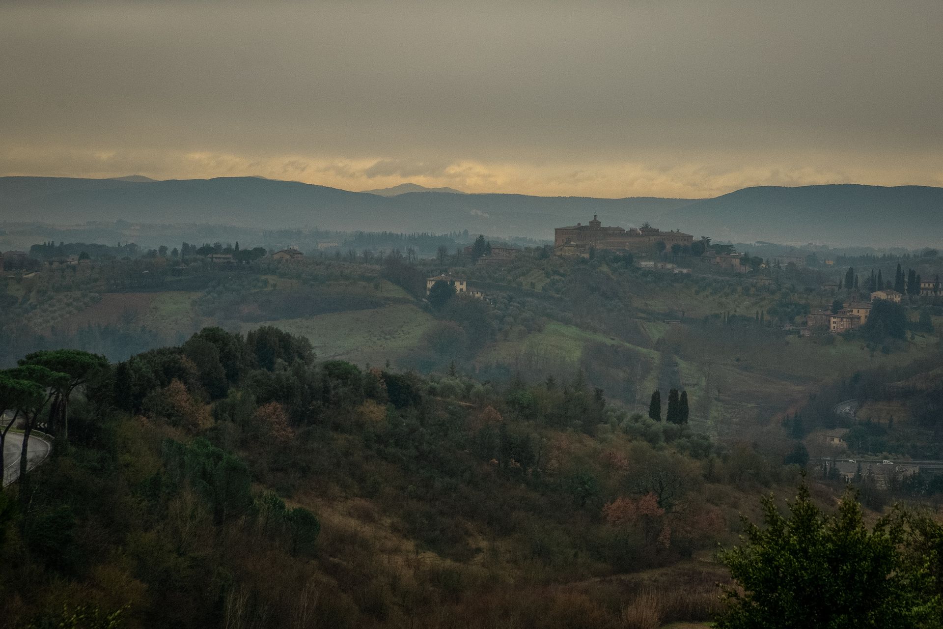 A serene view of the Italian countryside with rolling hills and distant mountains under a moody sky.