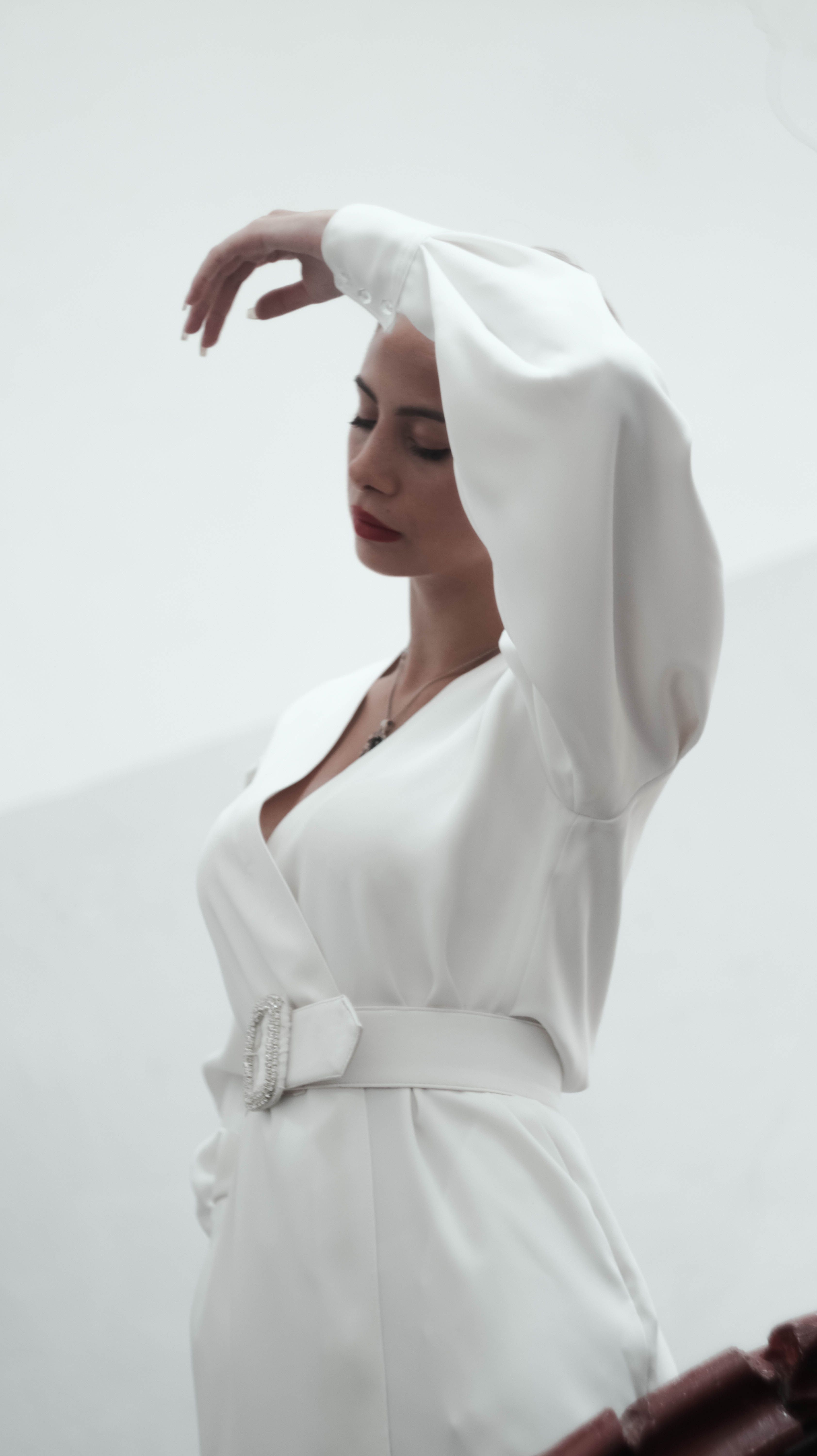 Elegant young female in vintage white dress standing with closed eyes and raised arm against  white wall