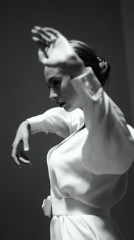 Black and white of graceful female with hairstyle in elegant white dress standing in dancing pose with raised arms against wall