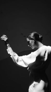Black and white of young female with hairstyle in white dress with puffy sleeves standing with raised arm against wall