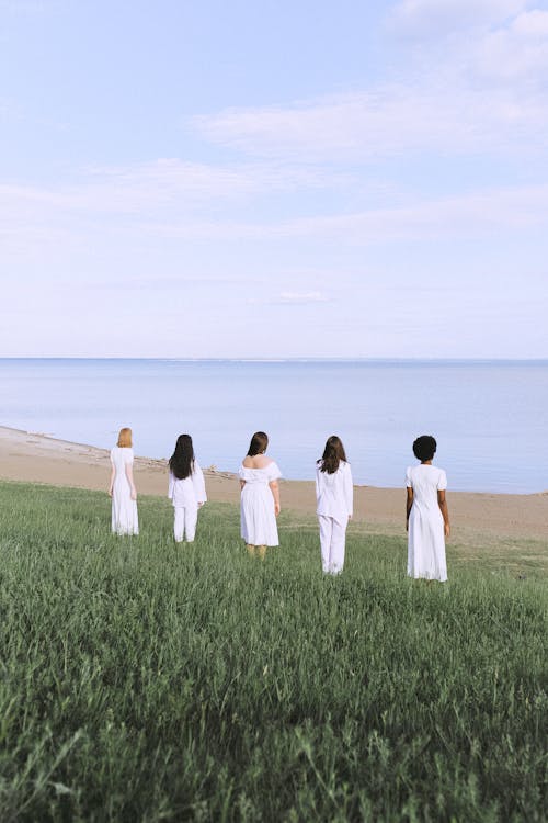 Women in White Dresses Standing on Green Grass Field Near Body of Water