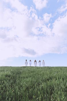Five women in white dresses standing in a green field under a cloudy blue sky.