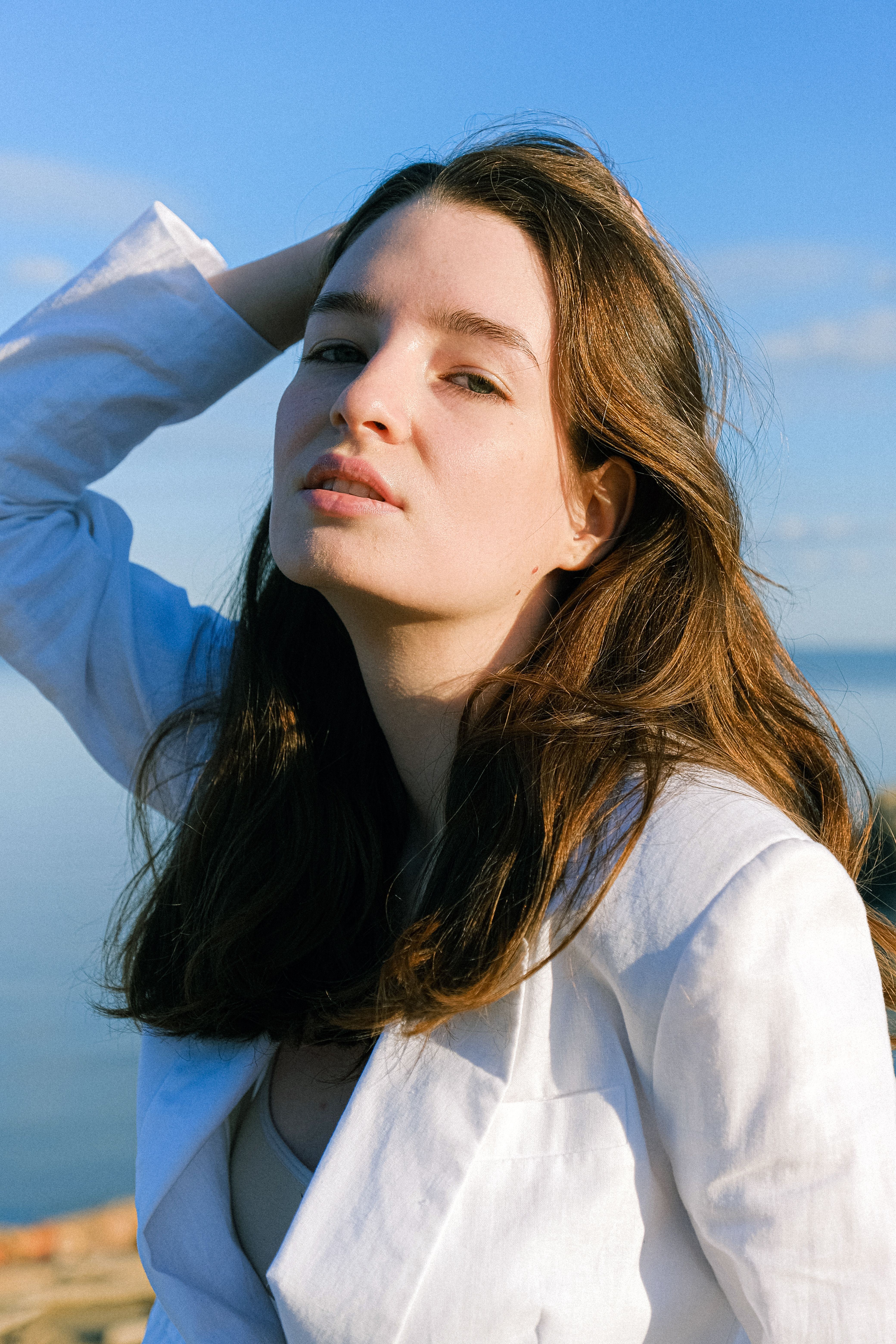 A young woman poses outdoors with her hand on her head against a clear blue sky.