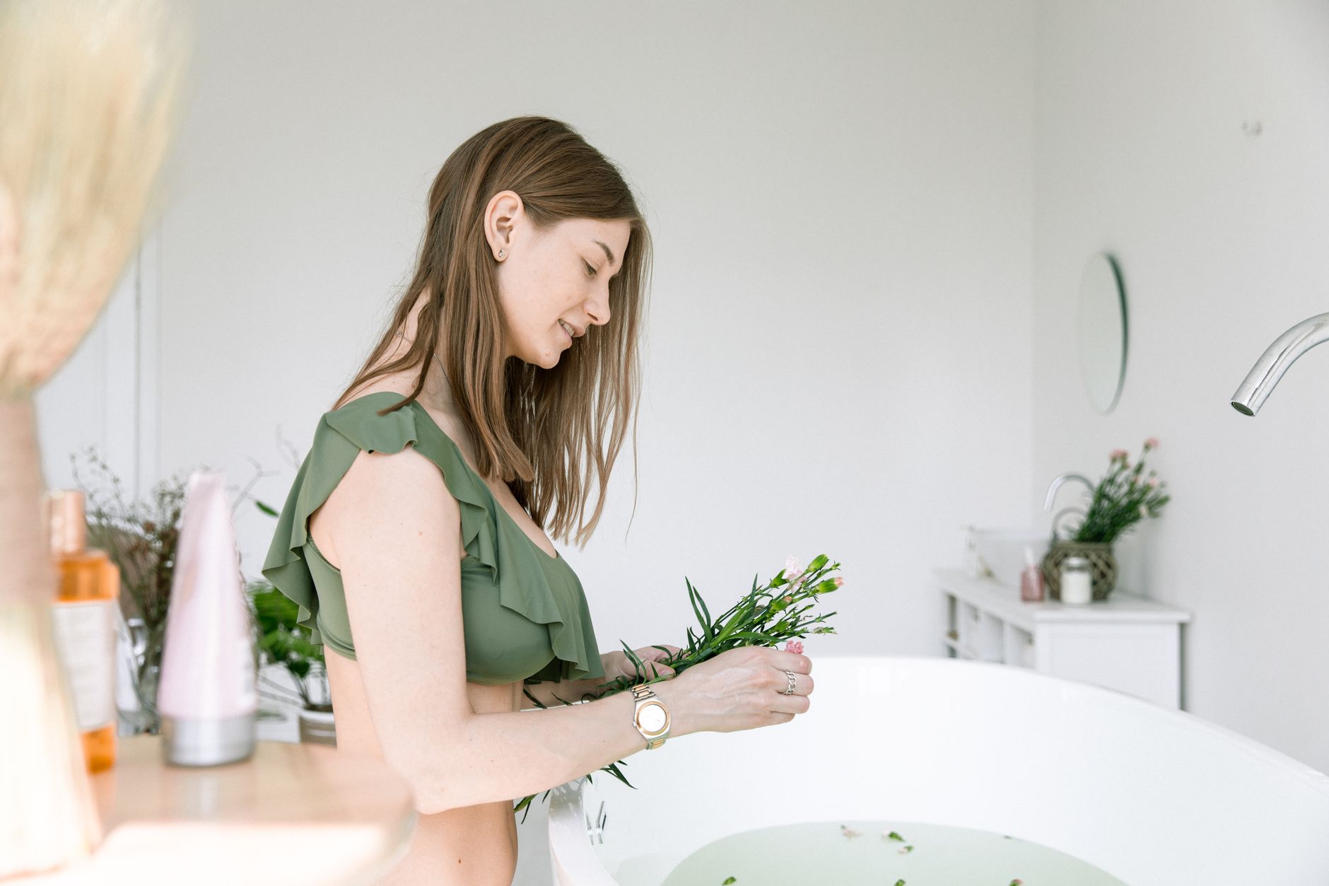 A woman in a bikini arranging flowers in a bathtub for a spa-like atmosphere indoors.