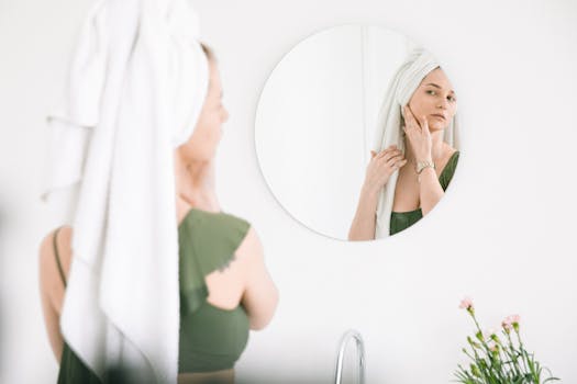 Woman in bathroom checking skin in mirror with towel on head.