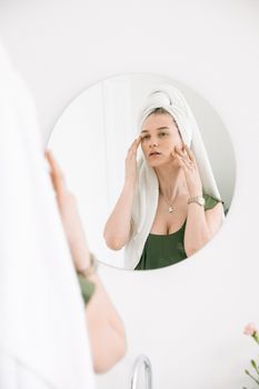 Woman in green top and head towel applies face massage in bathroom.