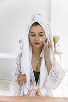 Woman in bathrobe applying face cream in a skincare routine indoors.