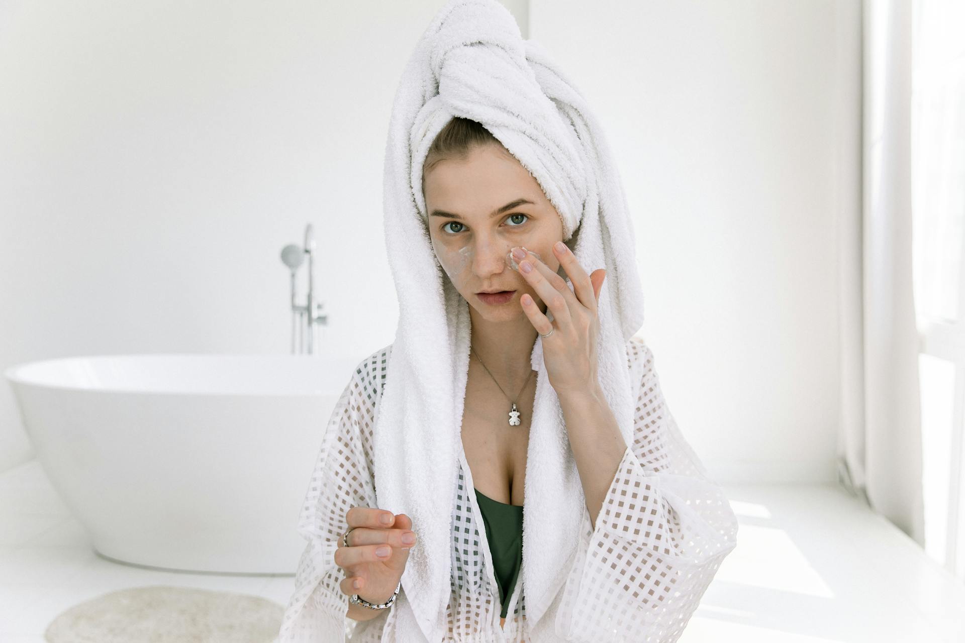 Woman in a bathrobe applying skincare cream in a bright spa-like setting.