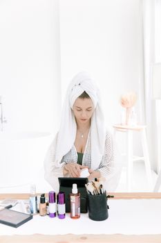 Woman in white bathrobe applying skincare products at a stylish indoor setup.