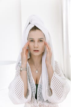 A woman in a bathrobe applies a facial mask, engaging in a relaxing skincare routine.