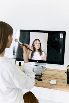 A woman follows a makeup tutorial on a computer screen during a video call, using a brush.