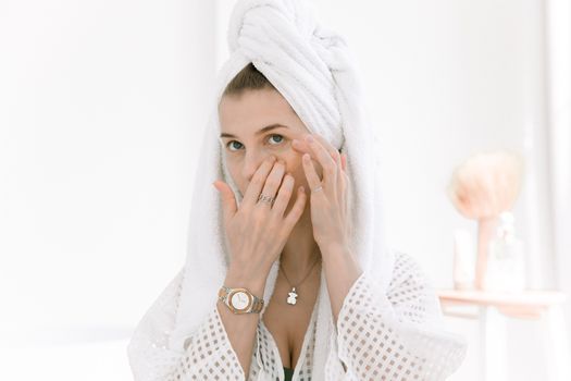 Woman in bathrobe applying skincare routine with a head towel indoors.
