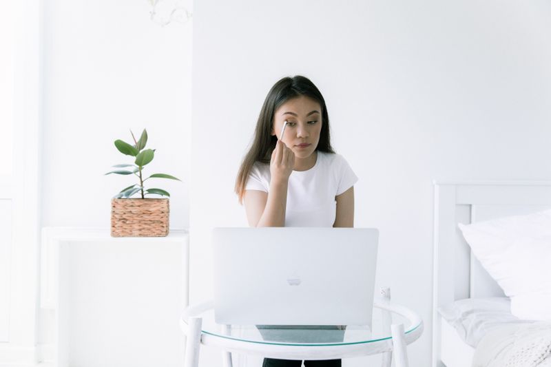 Young woman applies eye shadow while using a laptop in a minimalist indoor setting.