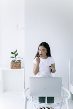 A woman applies lipstick indoors, using a laptop as a mirror, in a minimalistic setting.