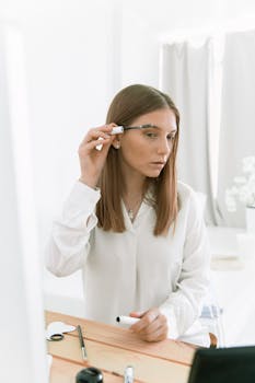Woman in white shirt applying makeup, highlighting her beauty routine in a bright and minimalistic setting.
