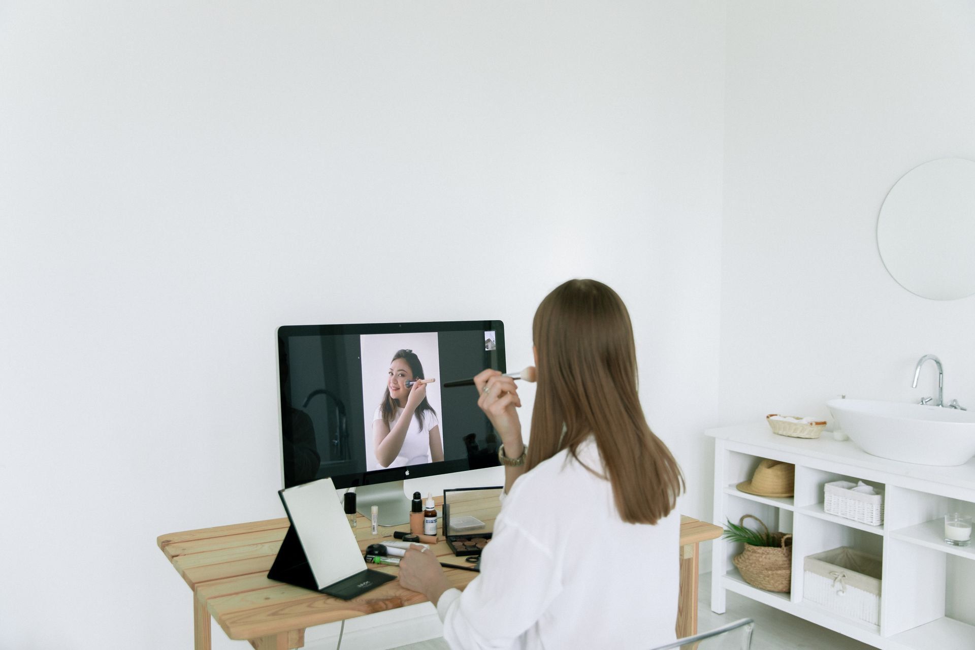 A woman uses a computer for a video call and applies makeup, showcasing modern beauty routines and technology.