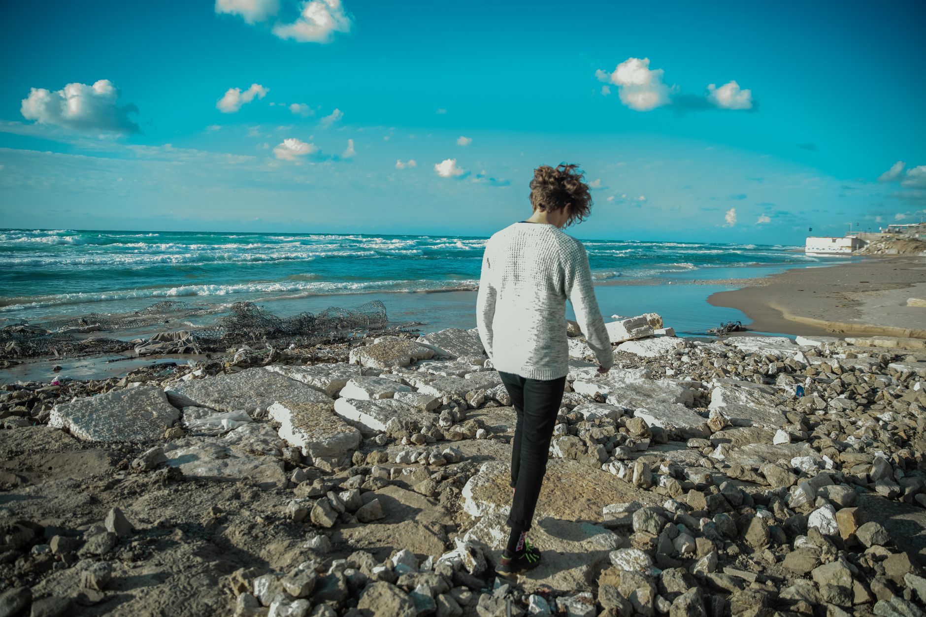 A young man with curly hair walks along a rocky beach in Israel.