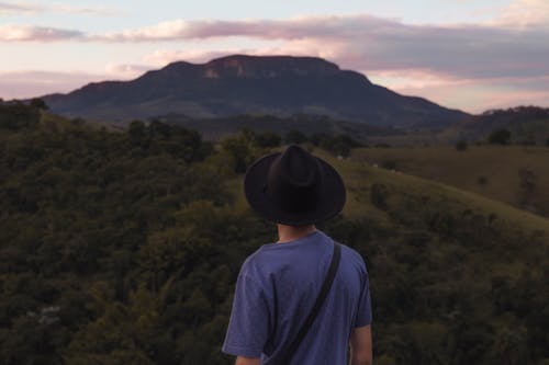 Man in Blue Shirt and Black Hat Facing Towards the Hills