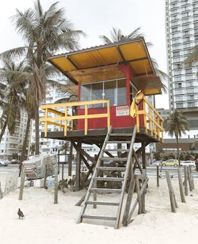 A bright lifeguard post stands on a sandy beach with palm trees in the background.