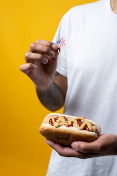 A person holding a classic hot dog with mustard and ketchup, waving a small American flag.