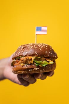 Close-up of hand holding a cheeseburger with an American flag on a bright yellow backdrop.