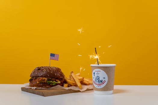 Celebrate Independence Day with a festive burger, fries, and sparkler on a yellow background.