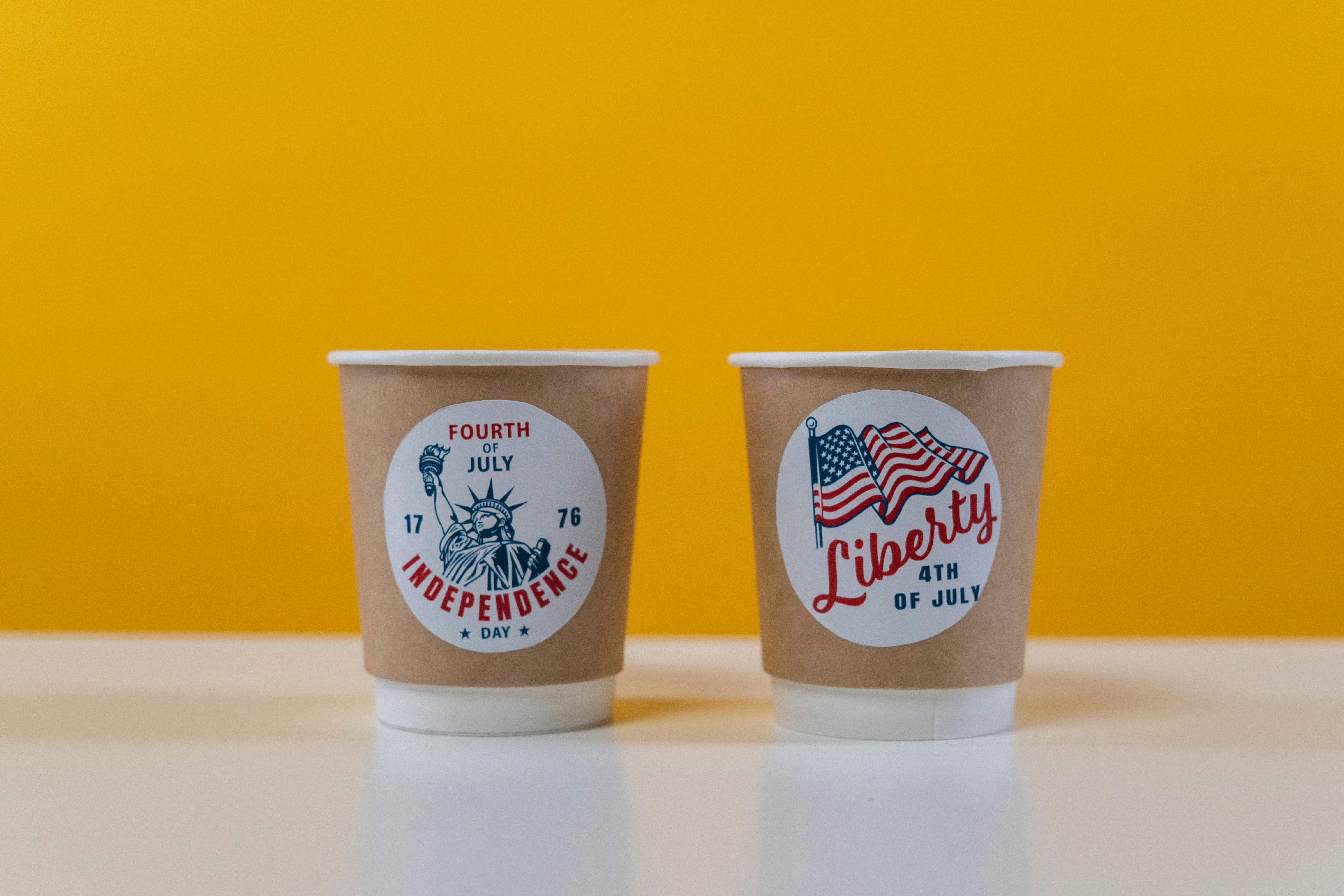Two coffee cups with Independence Day designs on a yellow background.