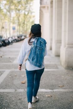 一名身穿牛仔夹克、头戴贝雷帽的女子，正走在城市的人行道上，留下一个背影
