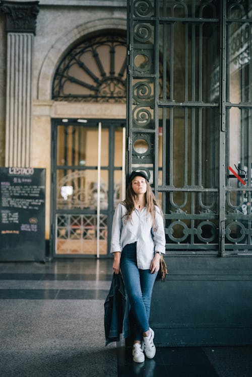 Donna In Camicia A Maniche Lunghe Bianca E Jeans In Denim Blu In Piedi Vicino Al Cancello Di Metallo Nero