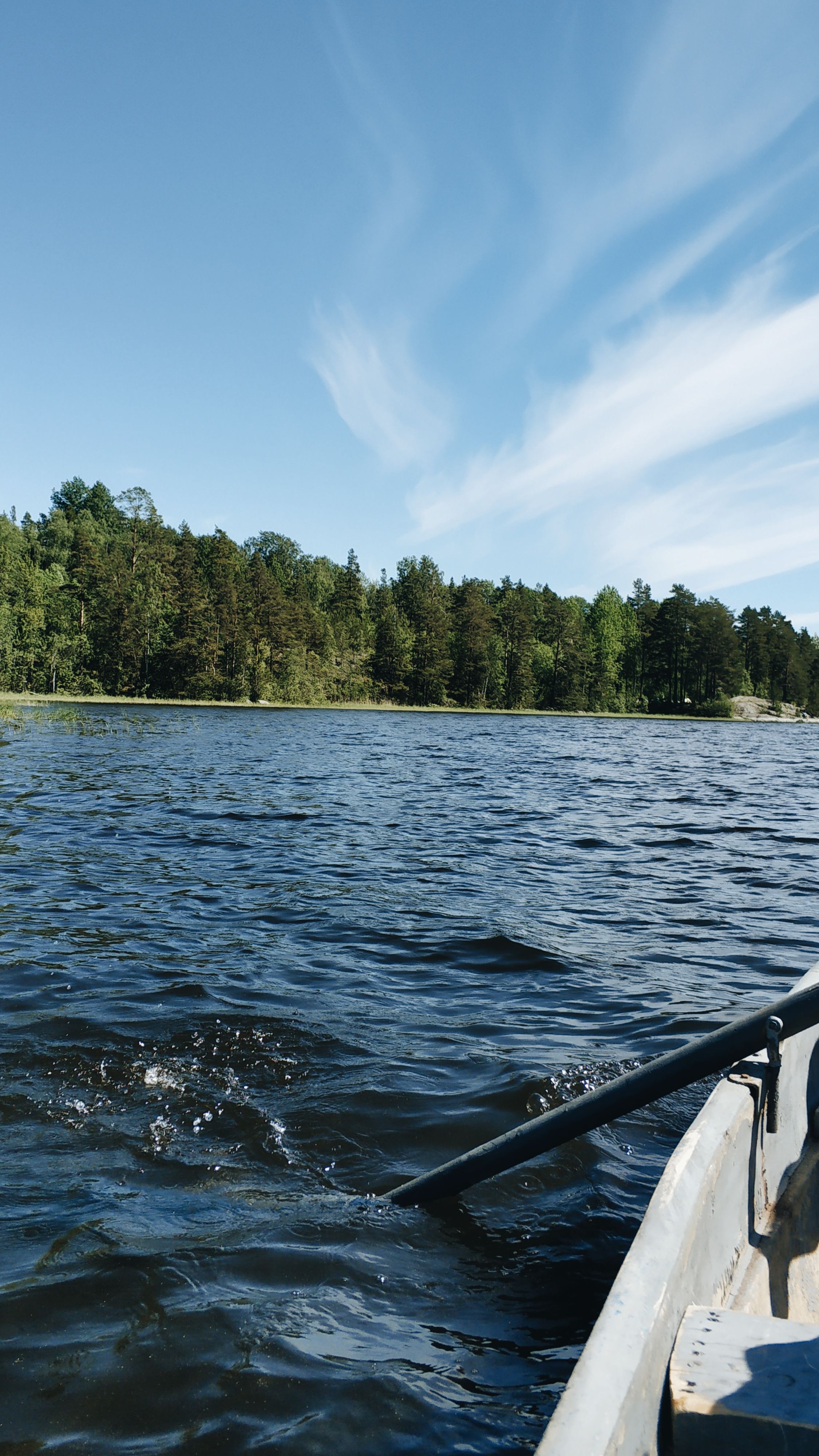 Wooden boat floating on surface of rippling lake surrounded by lush trees against blue sky in sunlight