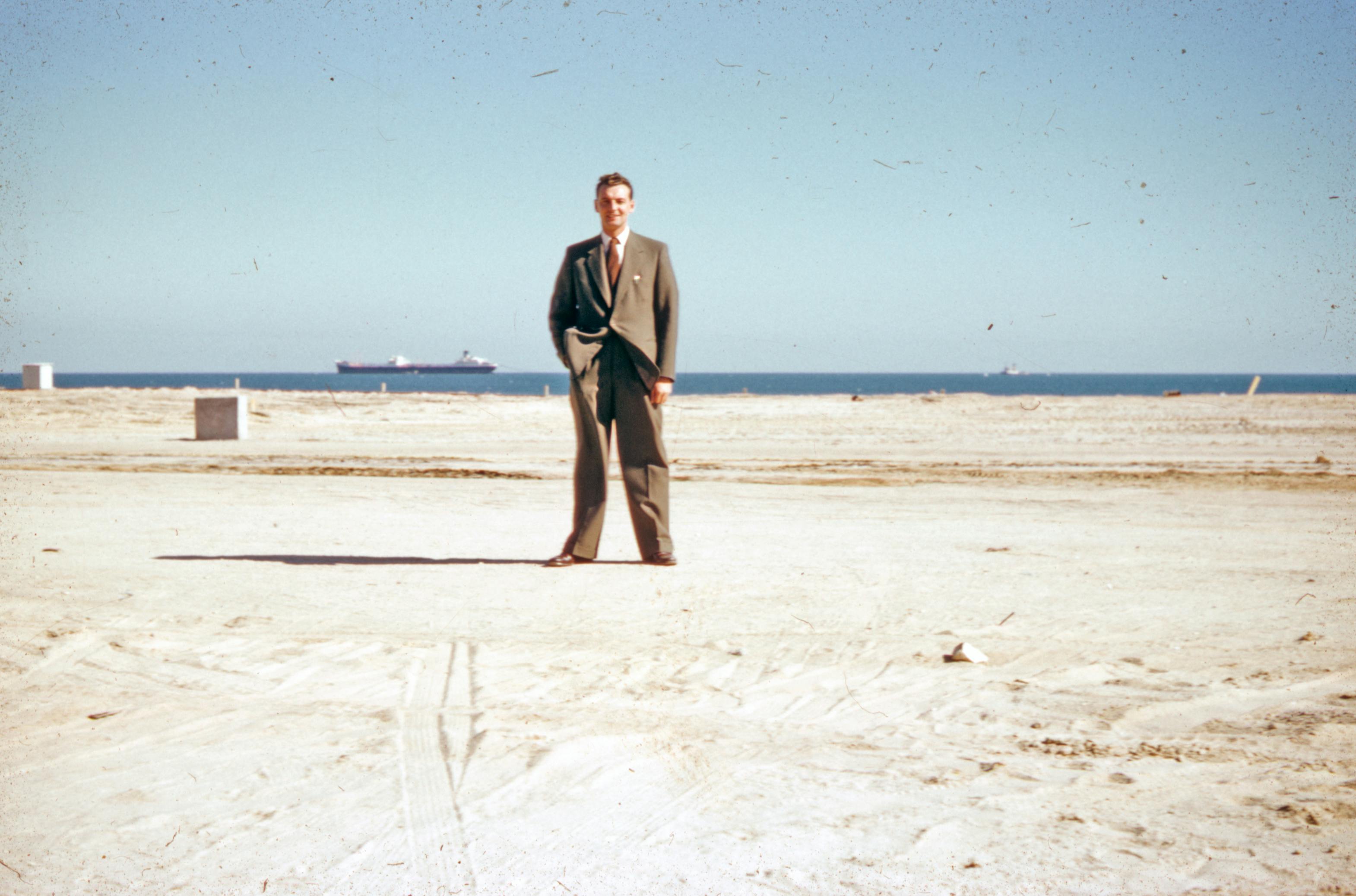 Vintage photo of a man in a suit standing on a sandy shoreline with the sea in the background.