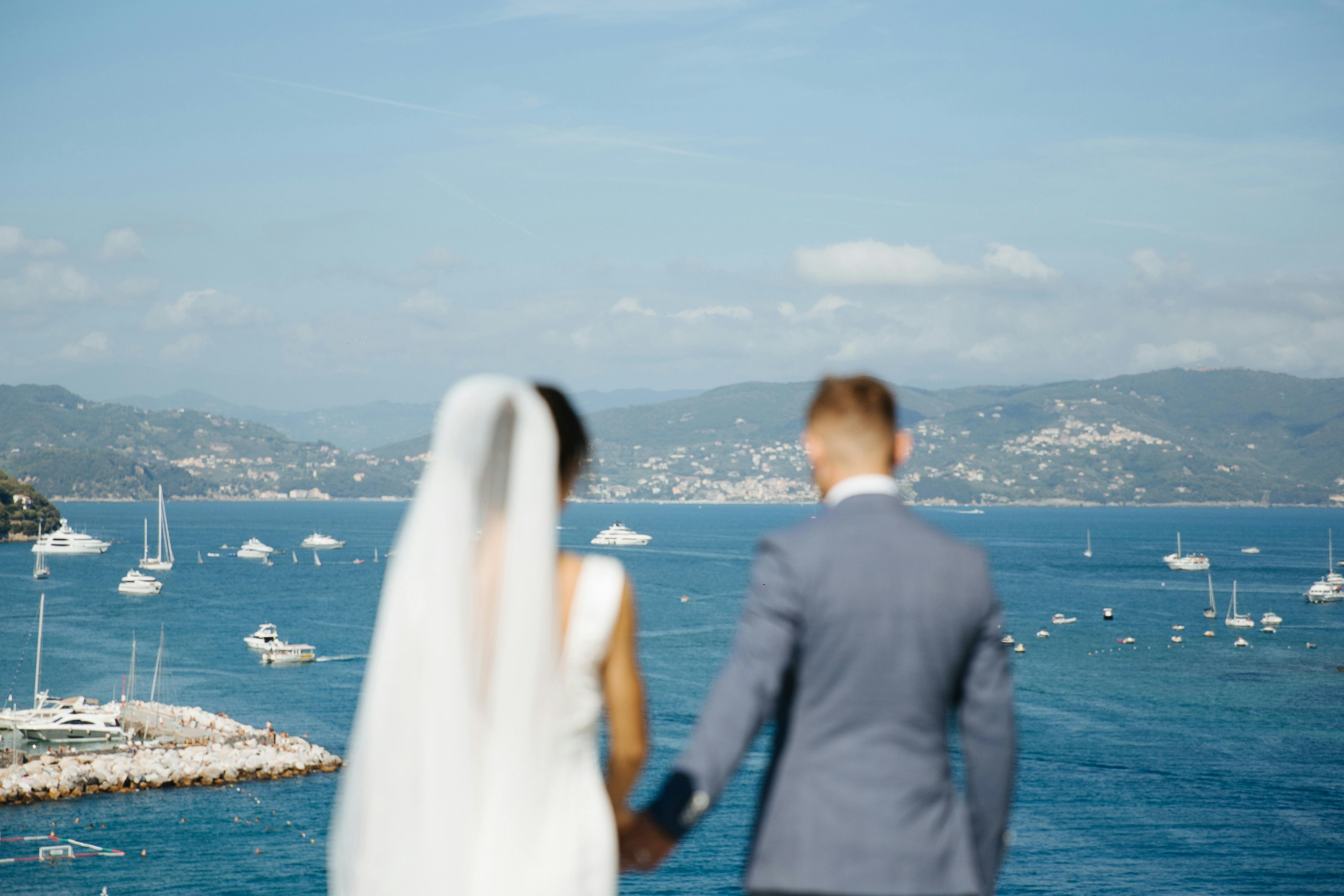 Newlyweds holding hands with scenic view of Porto Venere harbor, Italy.