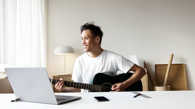 一名男子在家用笔记本电脑视频聊天，一边弹着原声吉他，一边享受音乐和联系