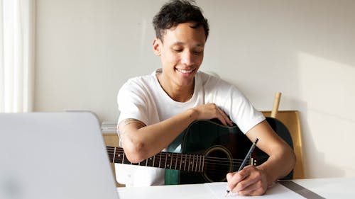 Man Holding Acoustic Guitar While Writing on a Paper