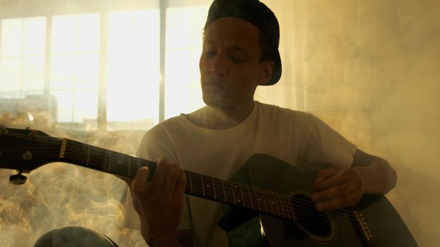 A young adult plays an acoustic guitar in a naturally lit, smoky indoor setting.