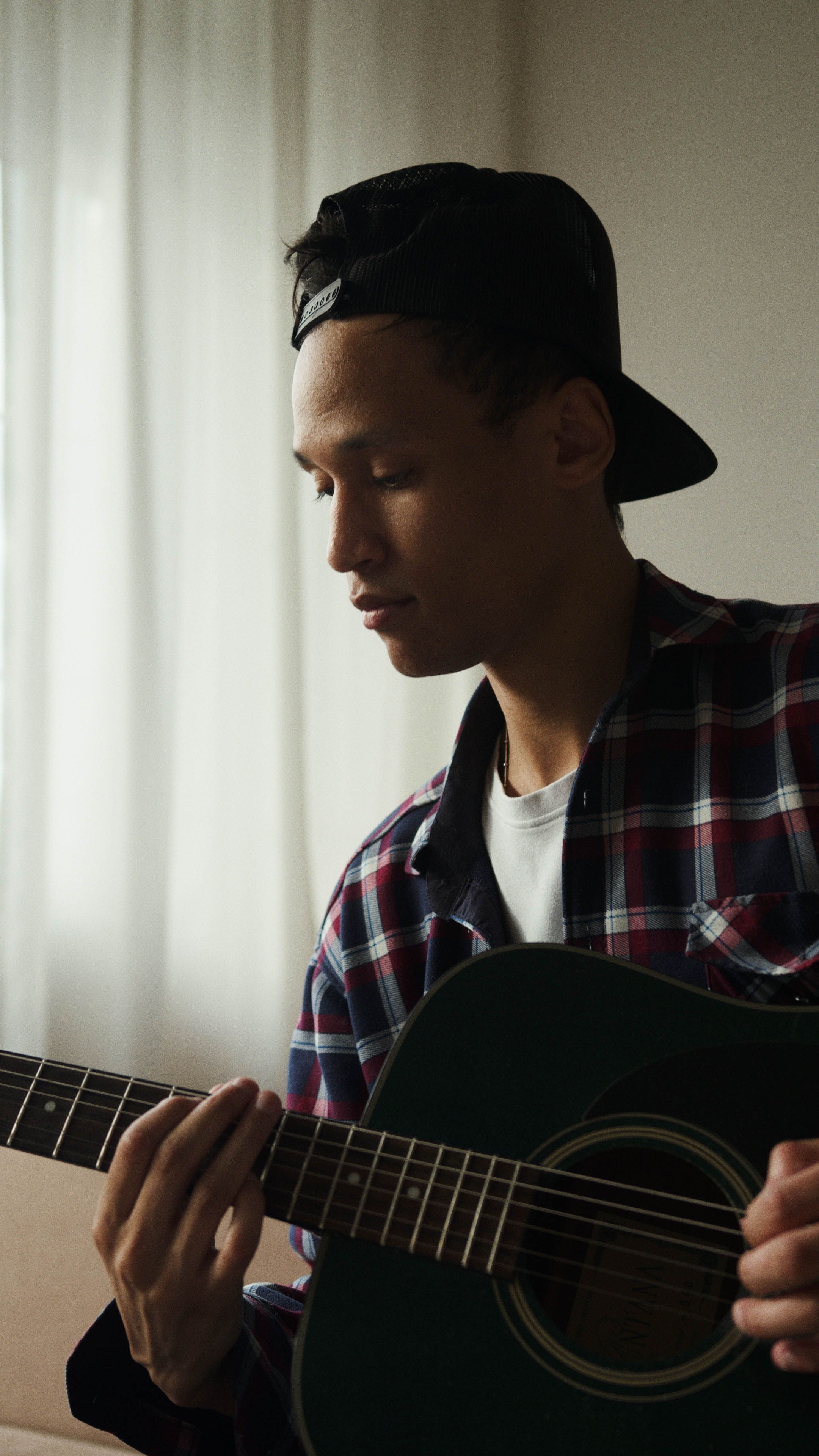 Young man in plaid shirt playing an acoustic guitar indoors. Captures musical focus.