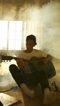 A young adult playing acoustic guitar in a smoky room with sunlight filtering through windows.