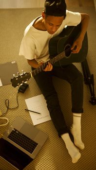 Young man learning guitar online, sitting on the floor with a laptop and notes.