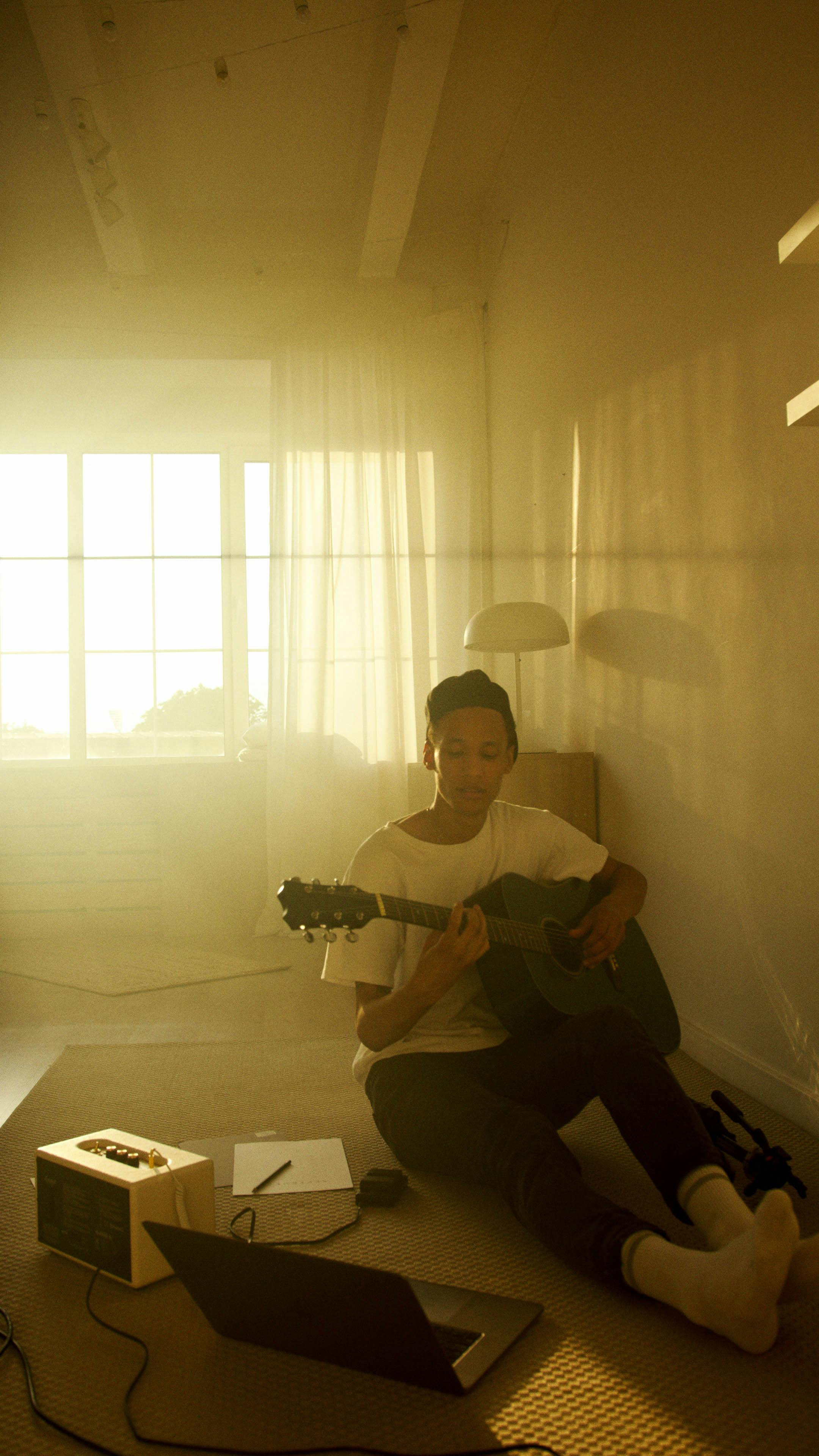 A young man sits on the floor playing guitar with sunlight streaming in through large windows.